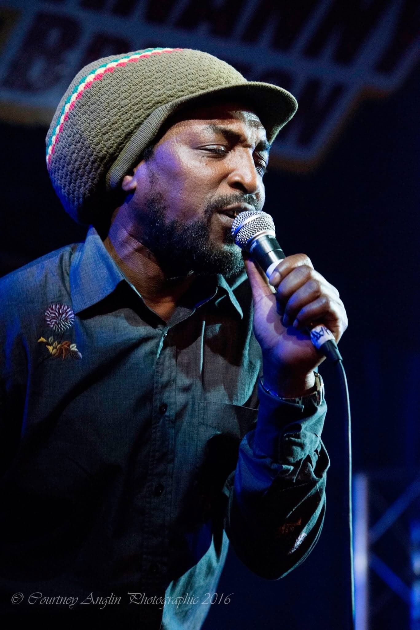 Kareem Shabazz singing into a handheld microphone under blue stage light at Hootananny Brixton, in a knitted cap