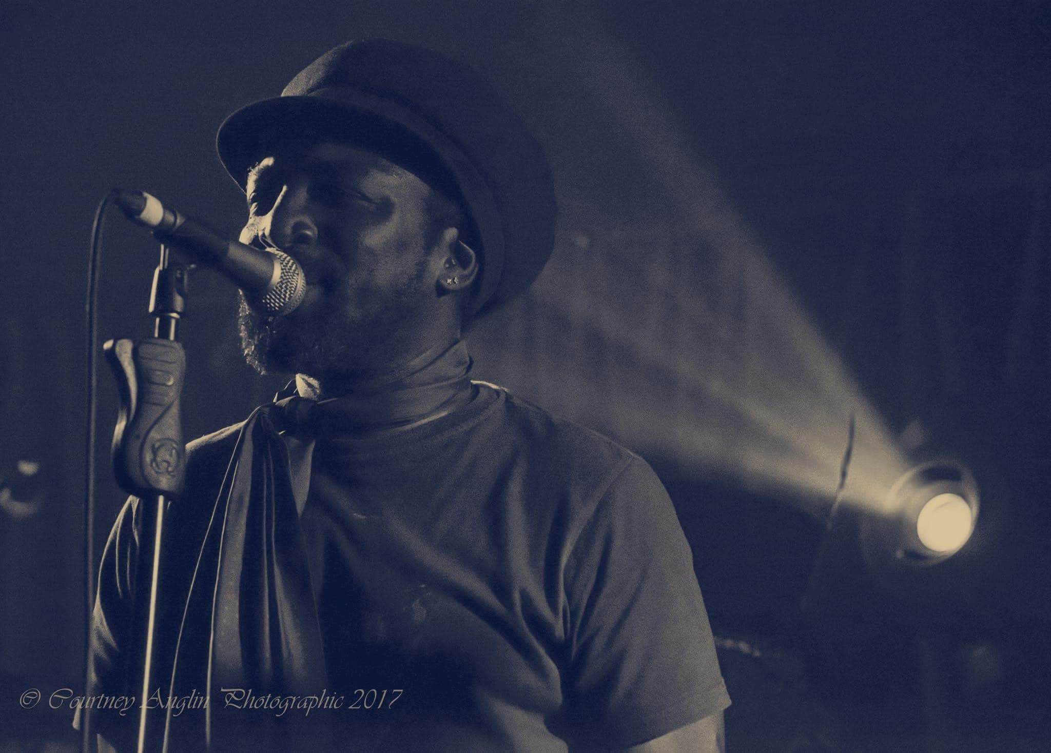 Kareem Shabazz singing at a microphone stand, a stage light beam cutting across behind him