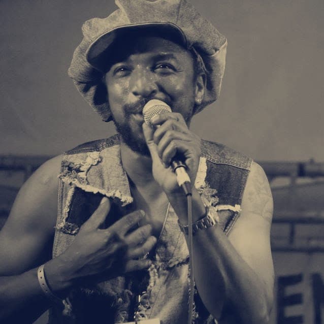 Kareem Shabazz singing with a hand on his chest in a patchwork denim waistcoat on an outdoor festival stage
